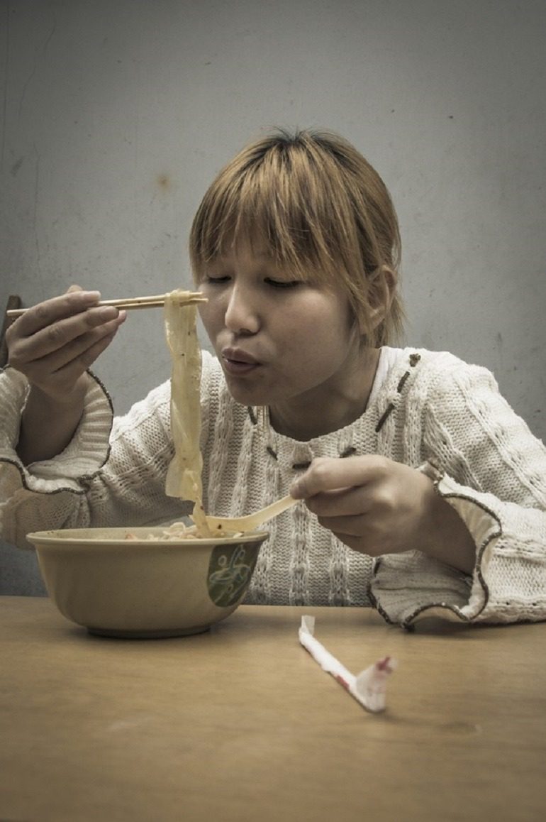 Image- A woman in a white sweater eats noodles in this street photography by Ron Gessel, take on ‘The Streets of Shanghai’