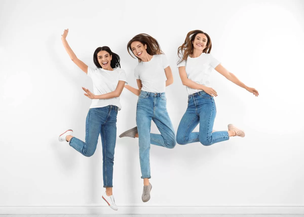 A group of three young women wearing sneakers, bootcut jeans, and white t-shirts smile as they jump into the air. Here are four shoes to wear with bootcut jeans in 2024 that will refresh their style and stay trendy.