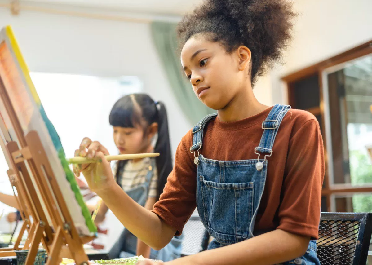 An African American child wearing overalls and a brown T-shirt painting on a large canvas with acrylic paints. This is why nurturing creativity and expression in young artists is rewarding.