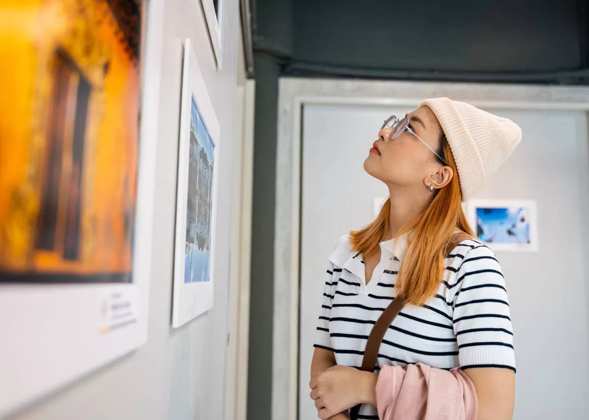 A young woman wearing glasses and a beanie studies a piece of artwork on the wall of an art gallery.