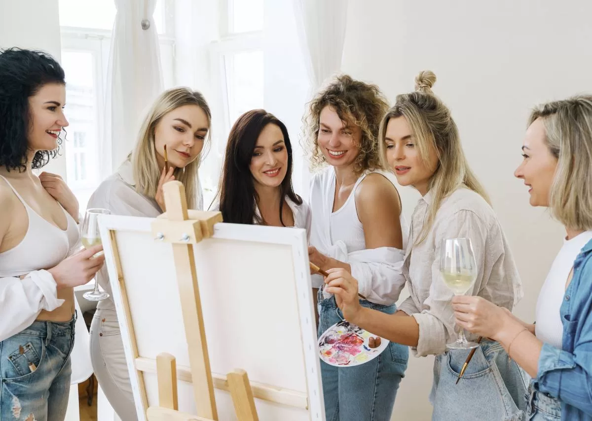 A group of women, with glasses of wine in hand, watch their friend showcase her artistic skills at an at-home art party.