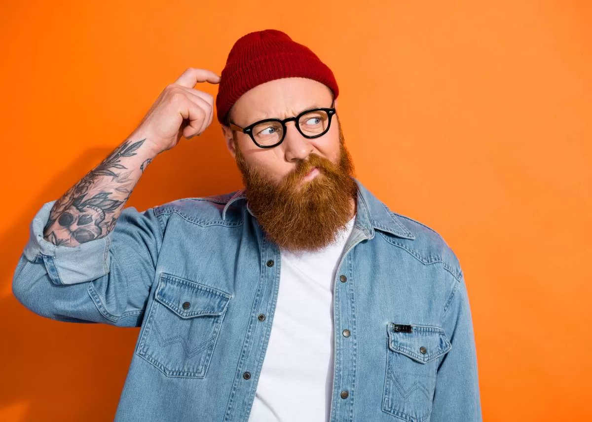 A long-bearded man with a tattooed arm, denim jacket, white shirt, glasses, and red beanie scratches his head as he ponders the common mistakes tattoo artists make in their career.