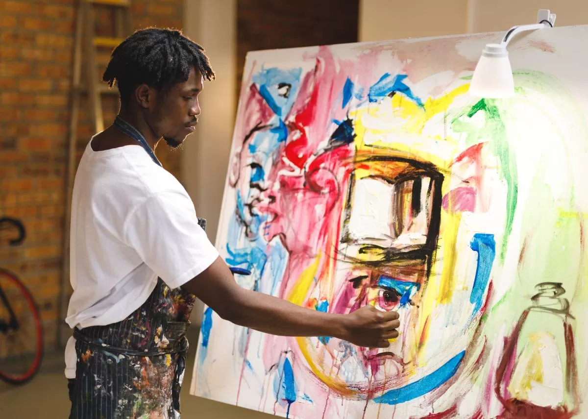A black male painter works on a large canvas in an art studio. He's creating a colorful, abstract piece. Like him, it is important to know how to sell your artwork without gallery representation.