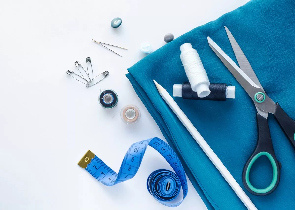 Sewing supplies, including blue fabric, scissors, a fabric pencil, a measuring tape, needles, thread, and safety pins.