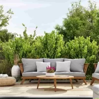 The sliding doors of a home are open. Light shines on a patio area with wooden furniture, gray cushions, and large shrubs. You can transform your-patio with expert exterior-design tips