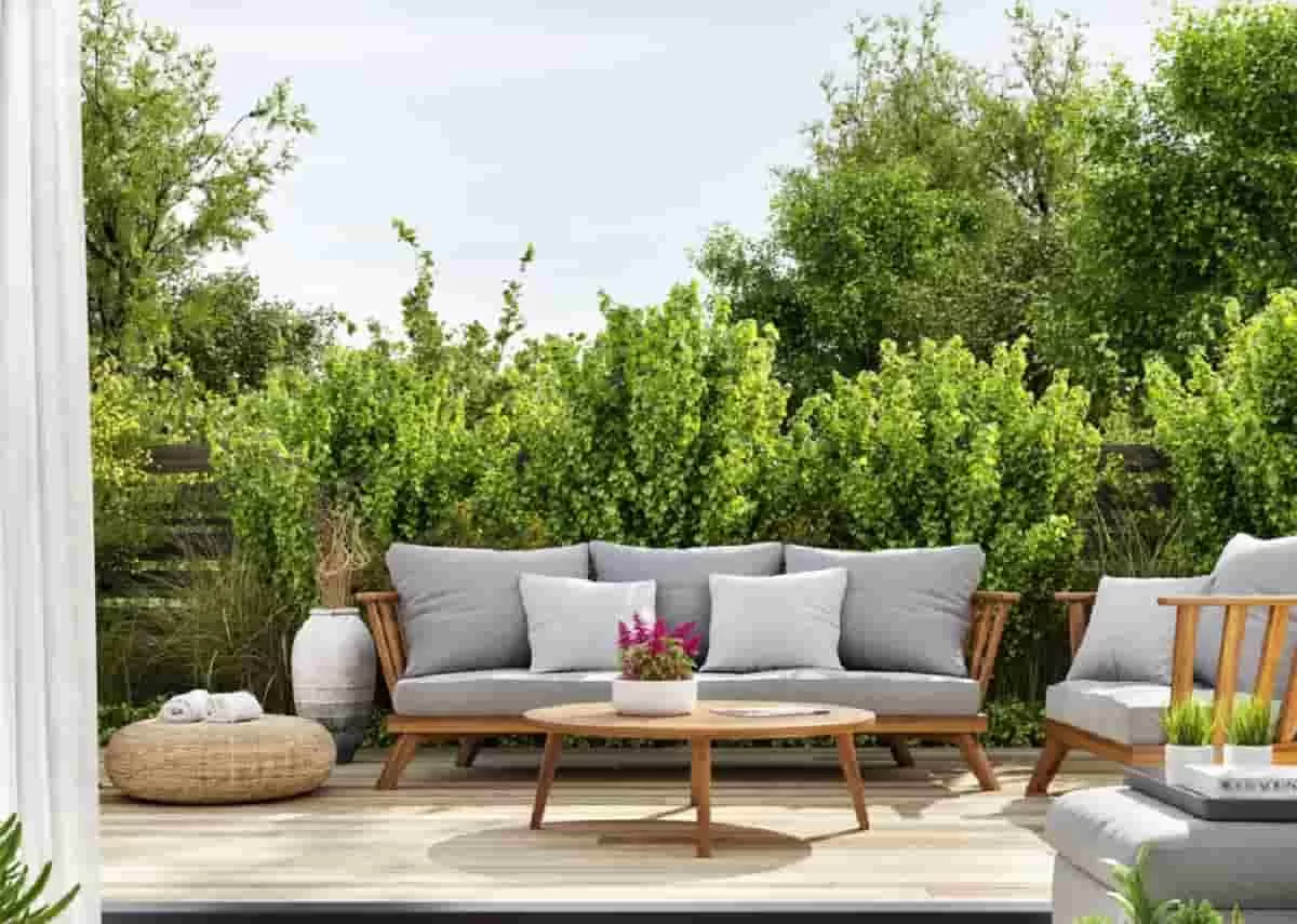 The sliding doors of a home are open. Light shines on a patio area with wooden furniture, gray cushions, and large shrubs. You can transform your-patio with expert exterior-design tips