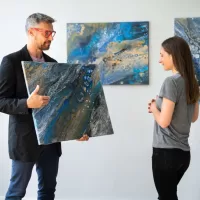 An artist holding an acrylic pour painting up for a potential customer to examine as he gestures to the work.