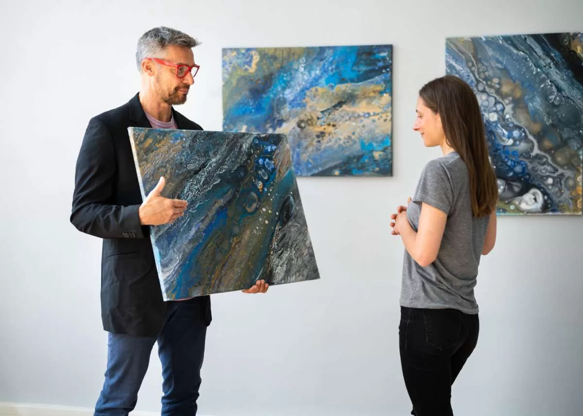 An artist holding an acrylic pour painting up for a potential customer to examine as he gestures to the work.