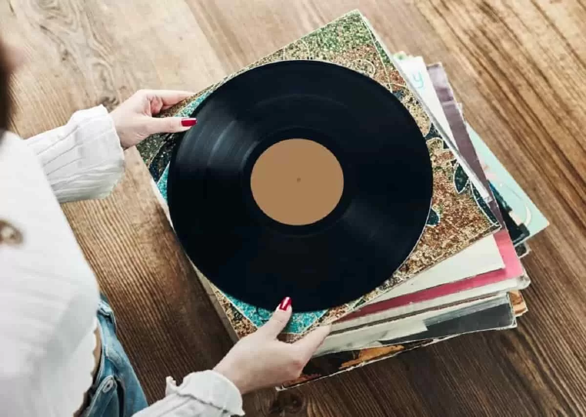 A woman holding a black vinyl record above a stack of vinyl records in their sleeves on a wooden table. This is an example of how to turn your vinyl collection into a gallery