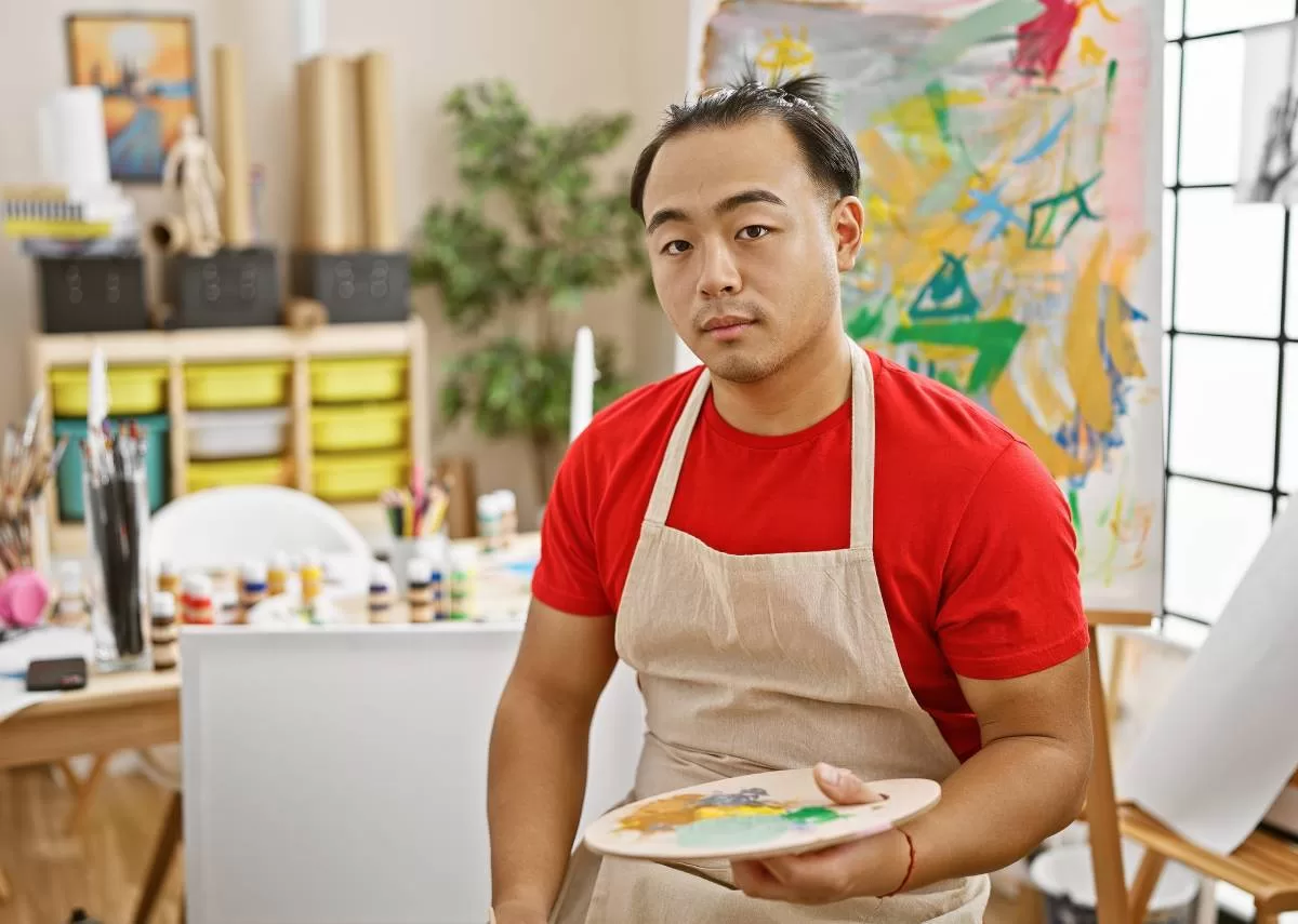 A serious artist holds a paint palette and sits in his art studio. Bottles of paint and brushes sit on a table behind him. To make art a career, it important to know how to showcase your artwork as an emerging artist