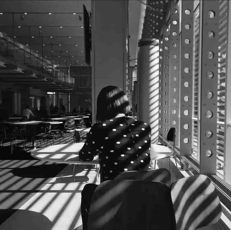 In this black & white photograph in Kathy Ryan, from Office Romance, a woman sits in a chair with her back to the camera. Flood of lights come from different direction casting multiple shadows across the floor and objects.