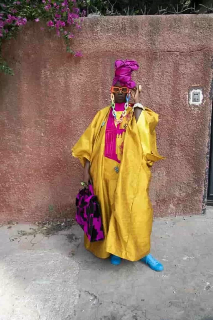 The street fashion photograph shows a woman dressed wearing a golden, well-designed, and embroidered gown. On her head is a towering purple headgear, which matches her purple and black handbag. She complements her appearance with a pair of blue shoes, purple glasses, and a watch with a white wristband. This is one of the photos in the street photography exhibition at the International Center of Photography.