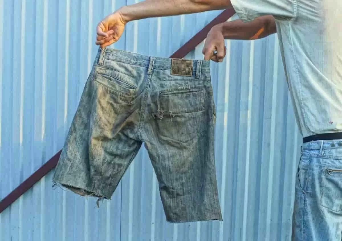 Someone holding a pair of distressed and deconstructed denim shorts off to their left side. The shorts are ripped and dirty.