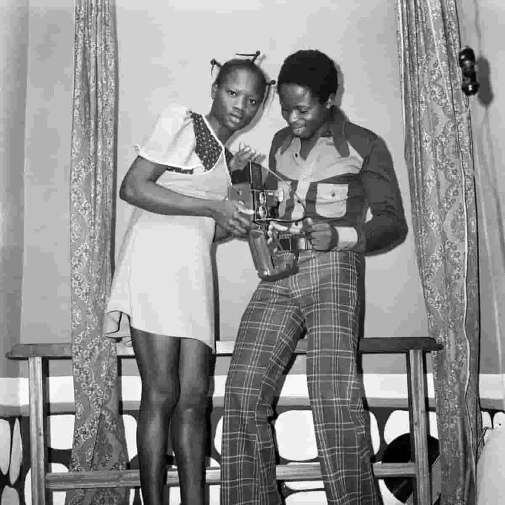 A man wearing a designer shirt and a bell-bottom pant poses with a woman wearing a minidress in a studio space with a damask curtain. While the man admires a camera, the woman gazes into the distances.