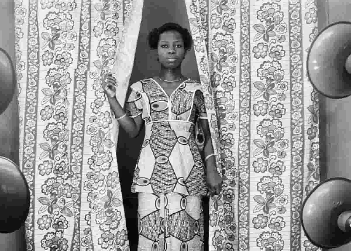 A beautiful woman dressed in ankara top and skirt poses between a flowery curtain and gazes into the distance. This is one the photographs in the Abi Morocco photos exhibition