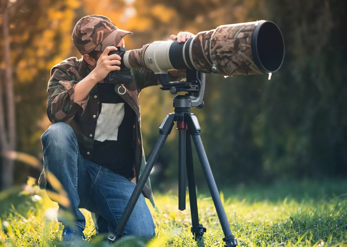 An outdoorsperson kneeling behind his large outdoor camera. The camera is facing into the woods and he's taking a photo. Taking a good outdoor photographs require the the must-have gear for an outdoor photoshoot