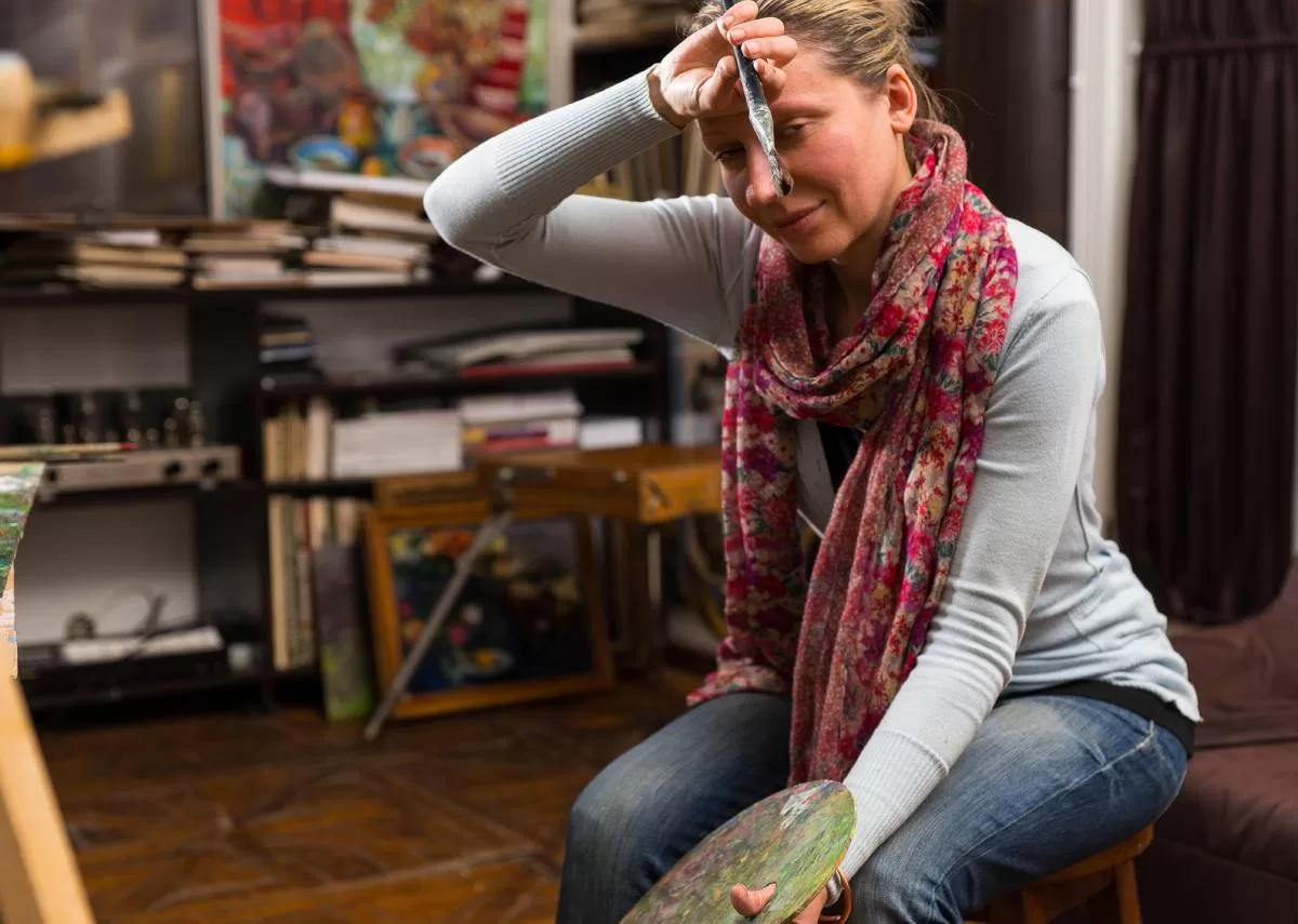A woman with a paintbrush in her hand wiping her forehead as she tiredly looks at the viewer. She has a paint palette in her hand. These tips can help her with her artist's block