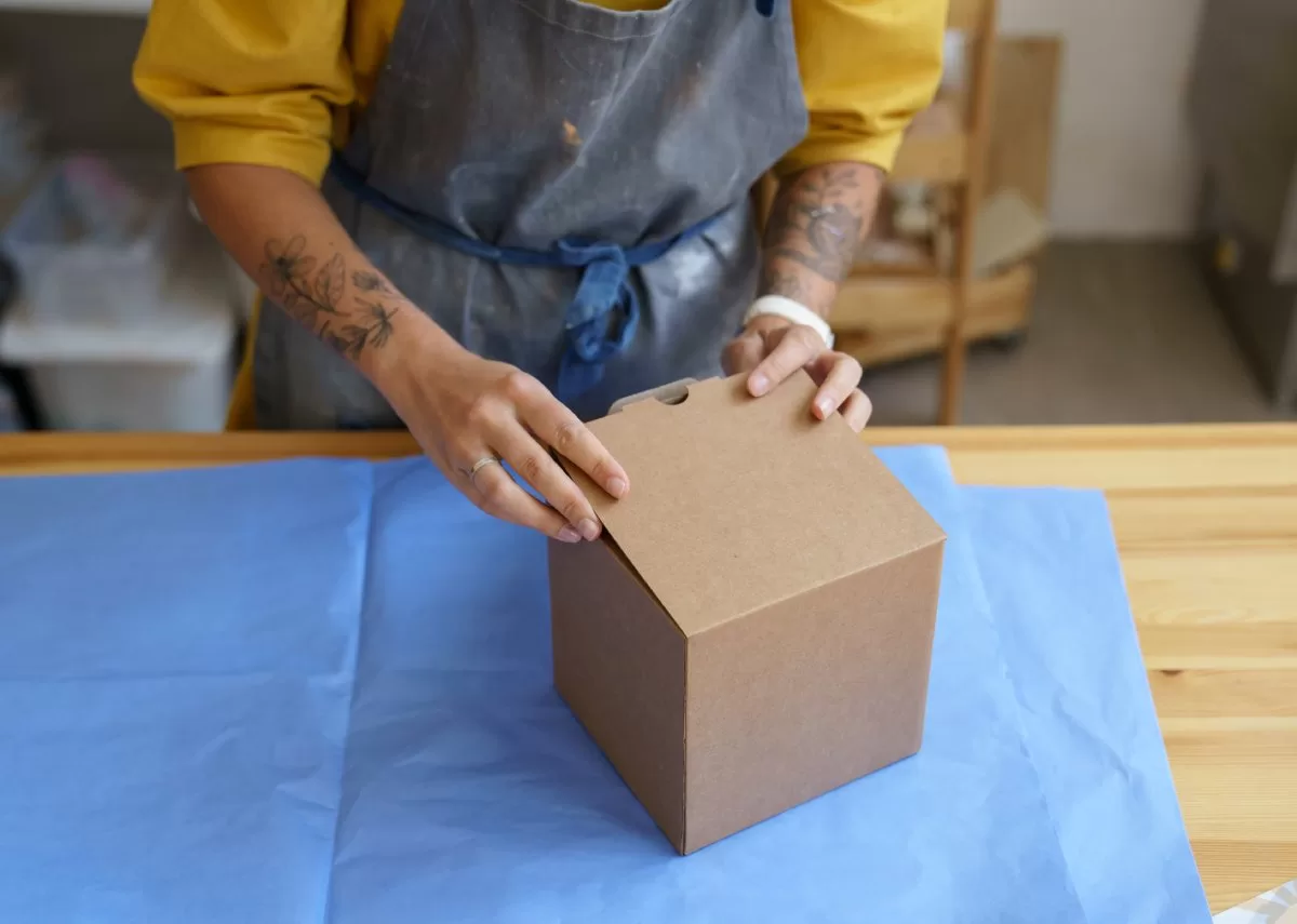 A woman wearing a smock covered in paint is closing the lid of a cardboard box on a worktable in a studio. It is important to avoid damaging your work when shipping fragile art pieces.