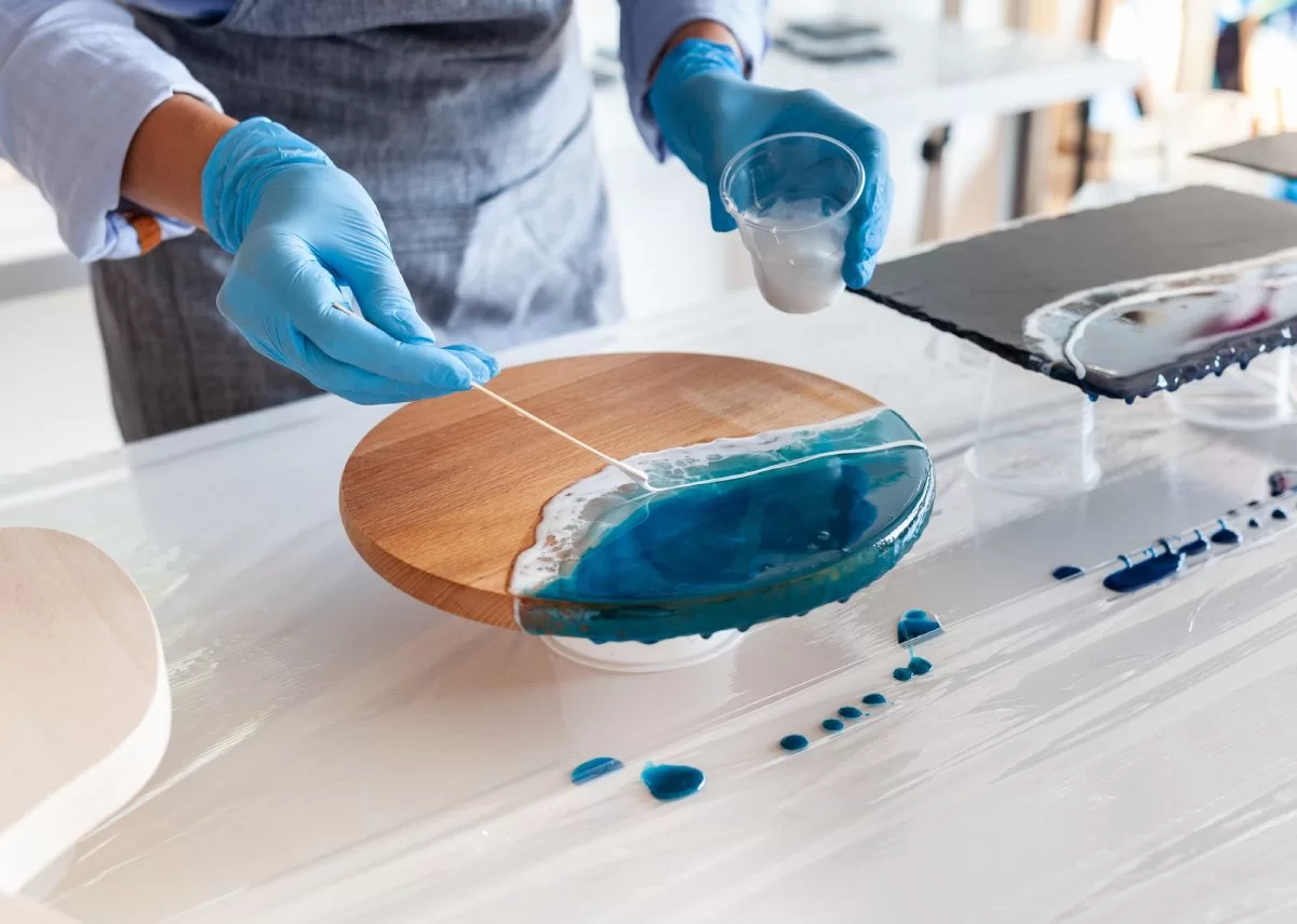 A close-up of someone doing epoxy art. The person wears gloves and uses a stick to place resin on the piece. Using proper tools and equipment are are tricks on how to achieve a flawless epoxy finish