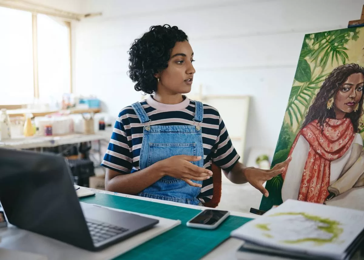 A young person sitting in front of an open laptop and gesturing to a large painting on display beside them. The question is Marketplaces or Stores: Which Is Best for Artists?