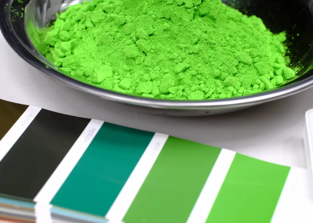 A bowl of green powder sits on a white table next to a paper with different green and brown color swatches. It is pertinent to select the best powder to ensure precision when powder coating your art piece