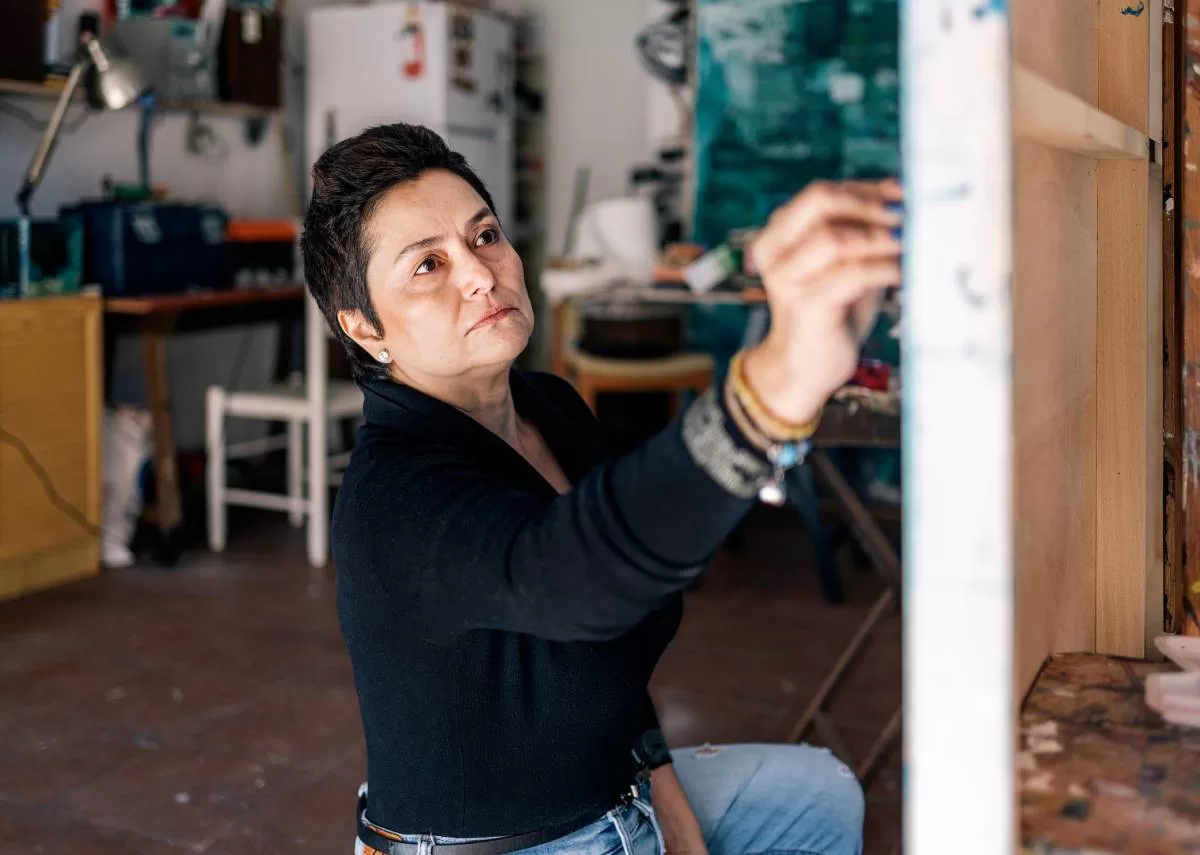 A woman raises a hand to color on a large canvas in front of her. Art supplies and furniture sit in the background. Proper organization is an important tip for building a backyard art studio.
