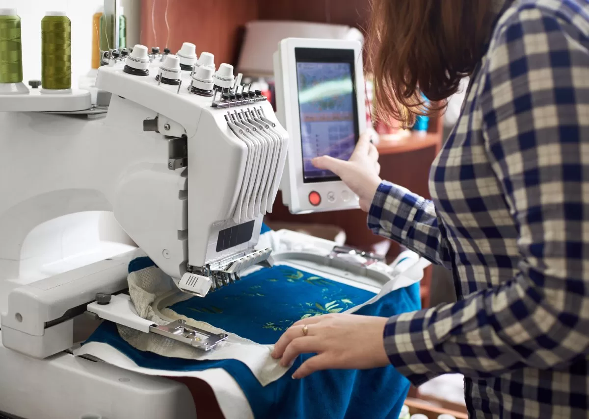 A close-up of a woman working on an embroidery machine with a multi-needle head creating a green floral pattern.