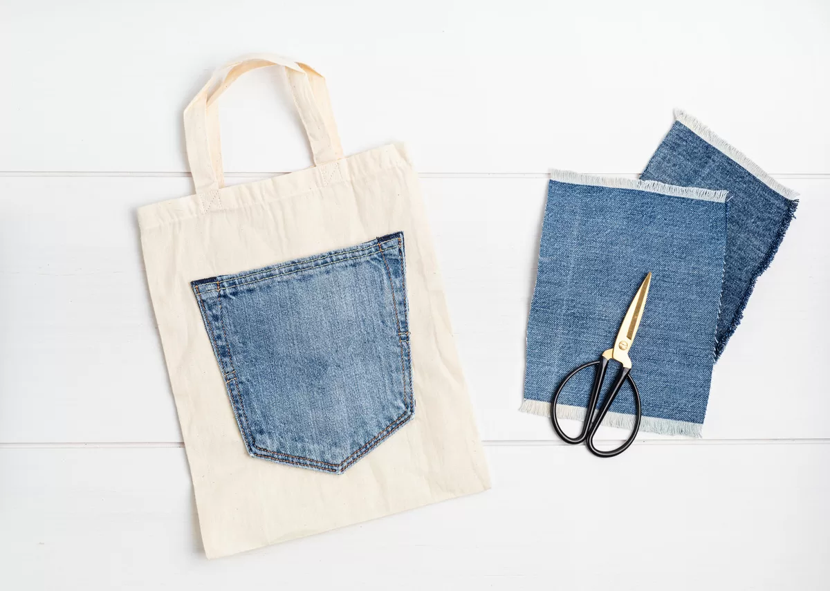 A white tote bag contains a jean pocket. Two denim squares and black and gold scissors sit next to the bag.