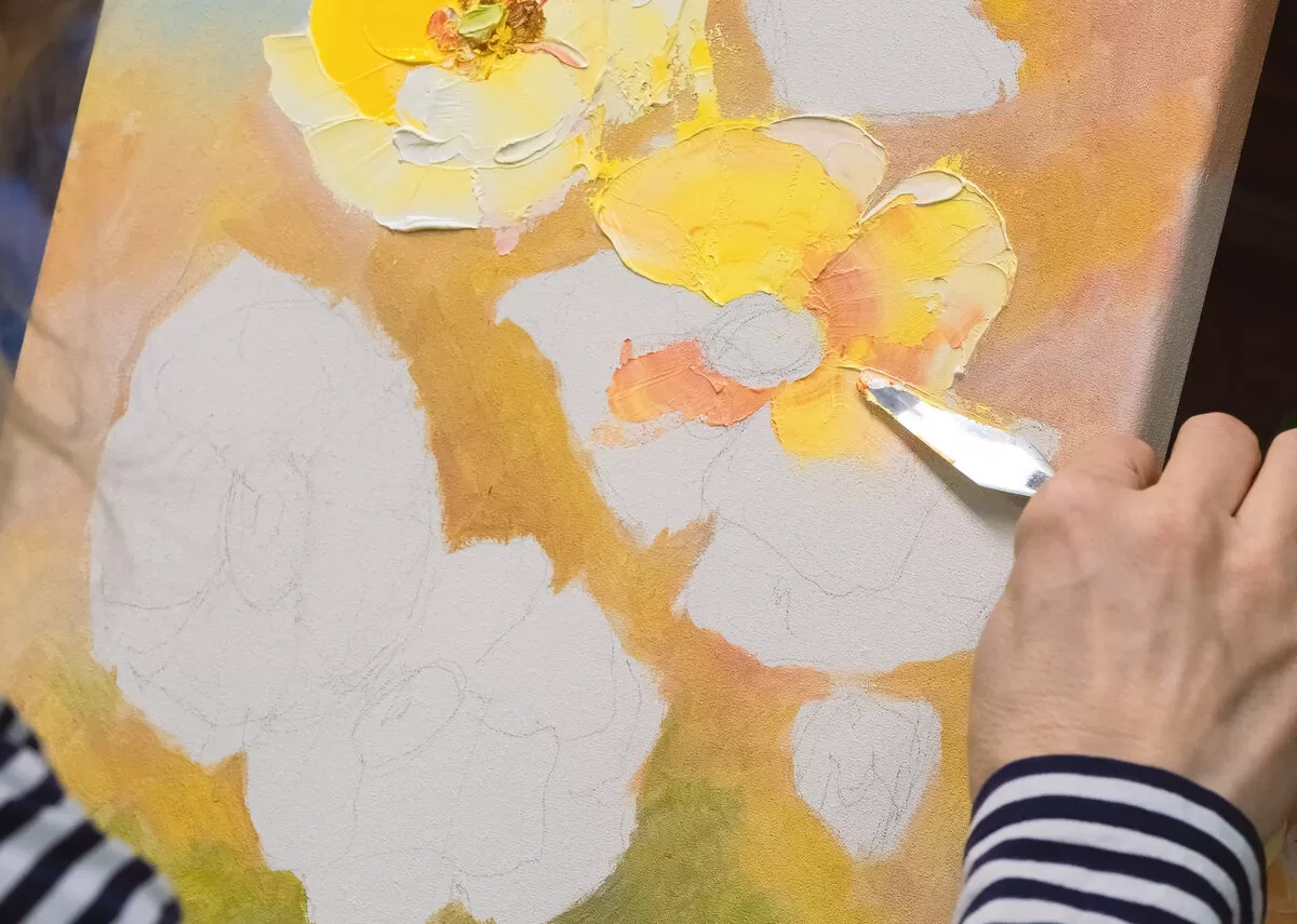 A close-up of a woman in a striped shirt using a palette knife to create petals on a pastel-colored flower painting on a canvas. Her style is one of the seven unique painting techniques you should try.