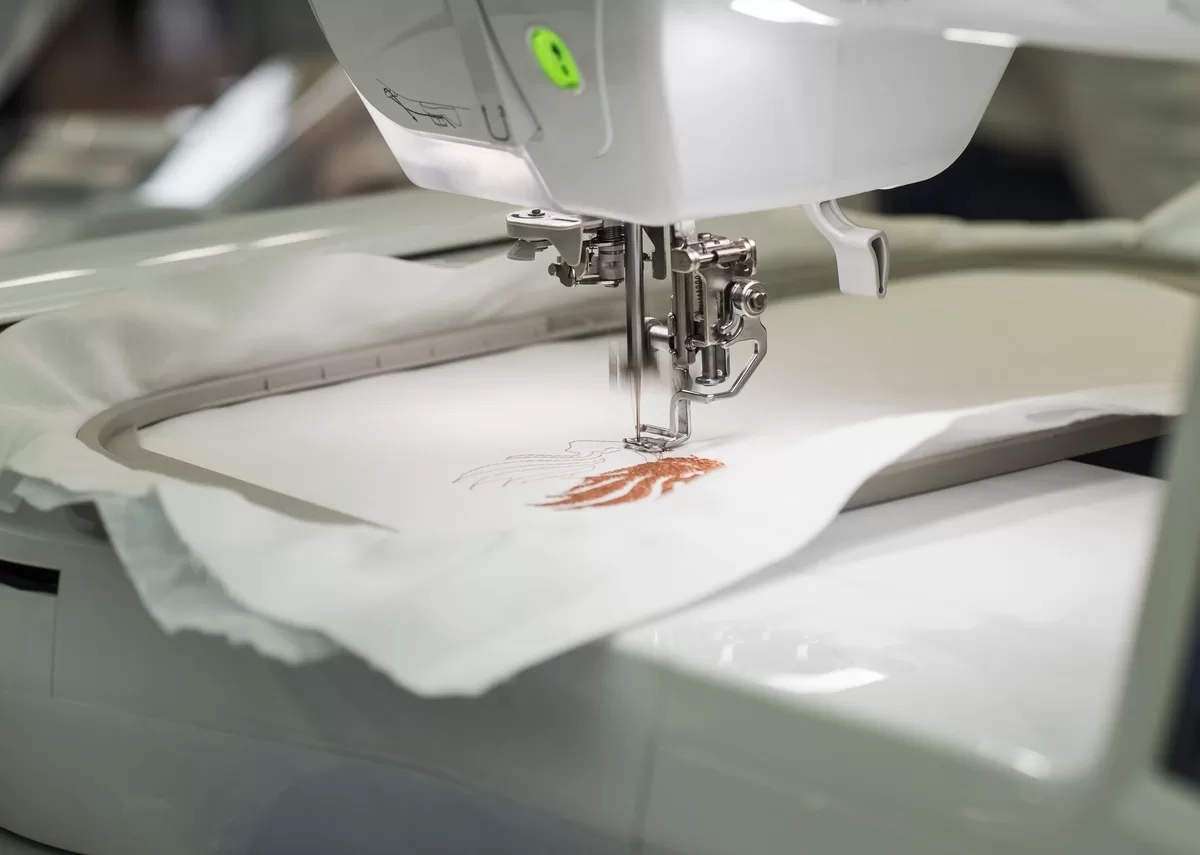 An embroidery machine embroidering a detailed pattern onto a piece of white fabric using copper-brown thread.