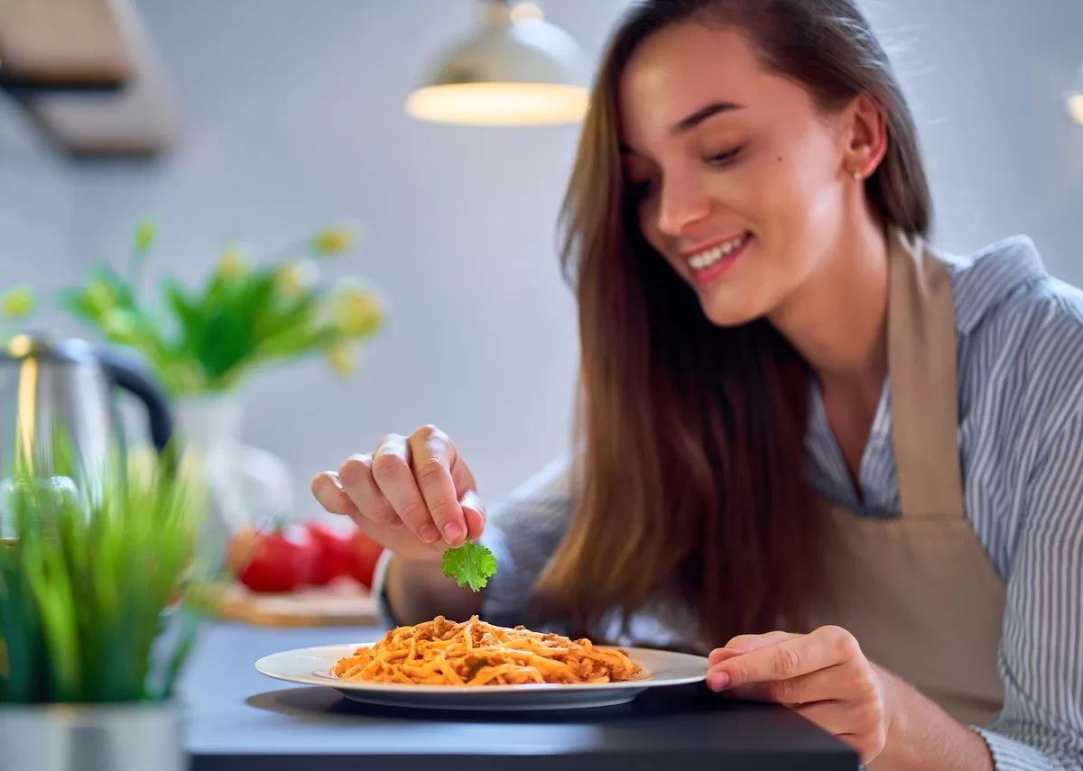 A close-up of a woman placing a parsley leaf on top of a plate of pasta. She's smiling and wearing an apron. Food is one of the Blank Canvas: Top Areas To Showcase Your Artistic Side