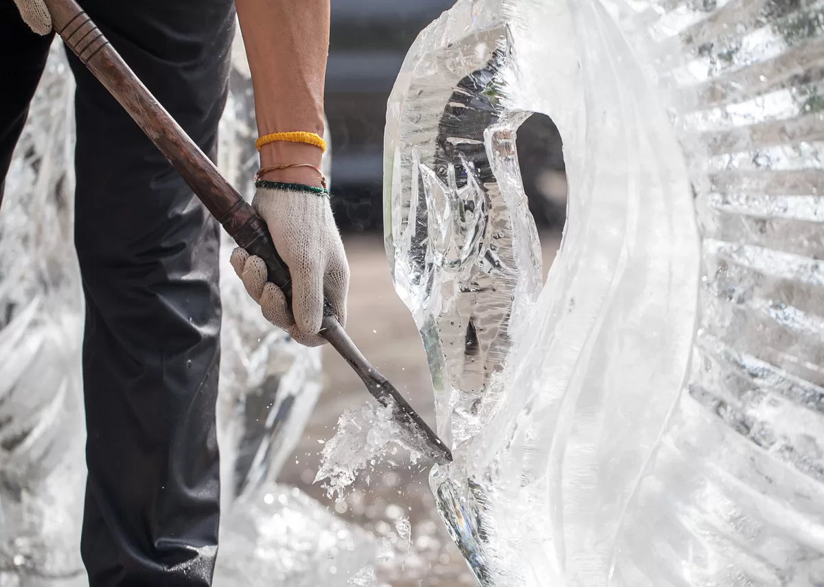 A pair of gloved hands chips ice off a swan sculpture’s beak, sending small ice shards into the air. Learn how ice sculptors keep their work cold and intact.