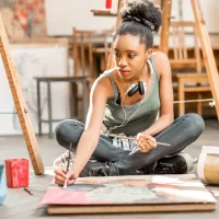 A young woman is listening to music as she paints on a canvas on the floor of her university's art studio.