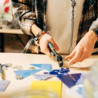 A close-up of a woman is in her art studio, putting together a glass mosaic with tweezers and a fusing tool.