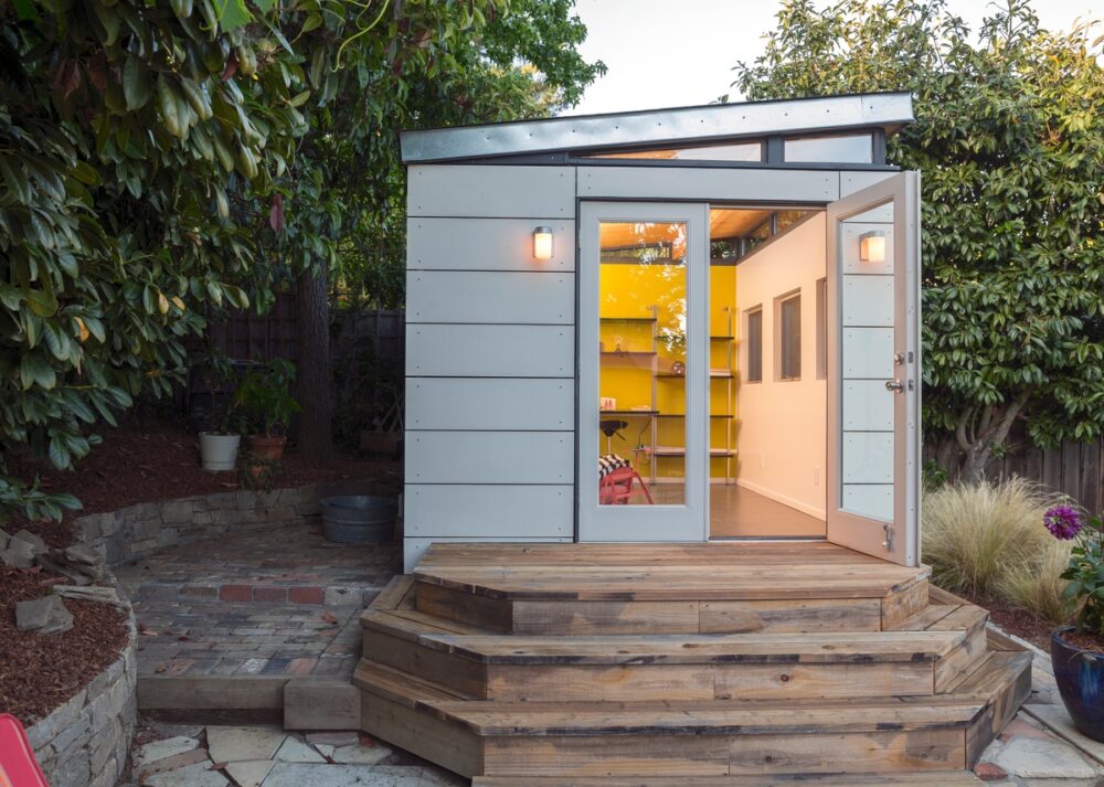A modern backyard studio with a slanted roof and open door sits on a wooden deck, surrounded by trees and greenery.