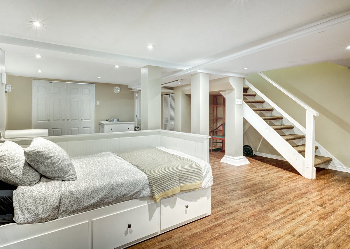 A bright finished basement with a white daybed, wood flooring, support columns, and a staircase to an upper level.