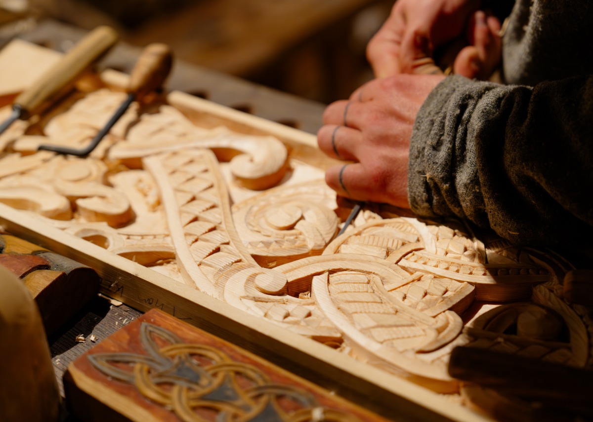 Carpenter carving detailed patterns into wood using hand tools, focusing closely on precise craftsmanship.