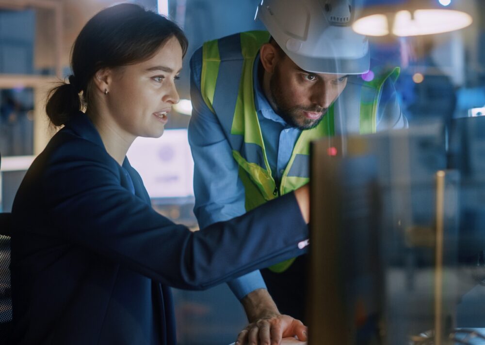 A female engineer and a male construction worker in safety gear are examining a digital blueprint on a tablet together. Collaboration is essential to designing industrial facilities for flexibility