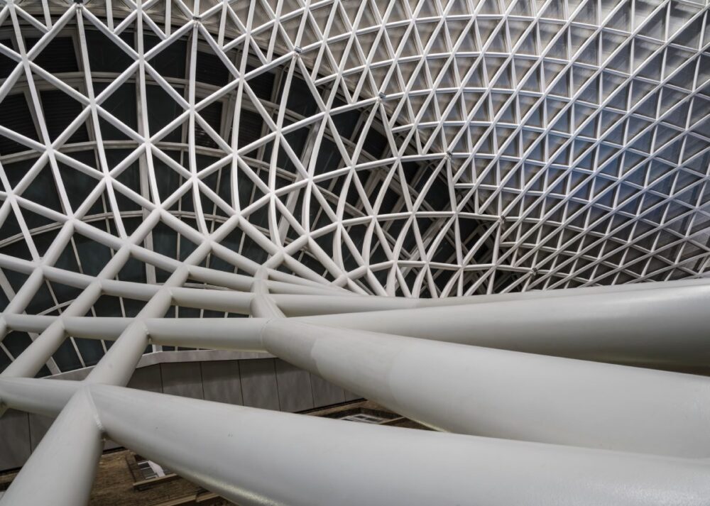 A white geometric lattice facade with interwoven metal beams forming a complex pattern over a glass structure.