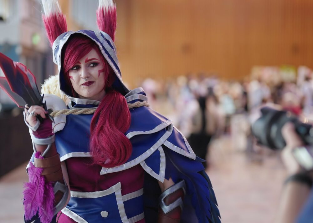 A woman in a blue cape and hood poses for a photo in a large convention hall with a crowd visible in the background.