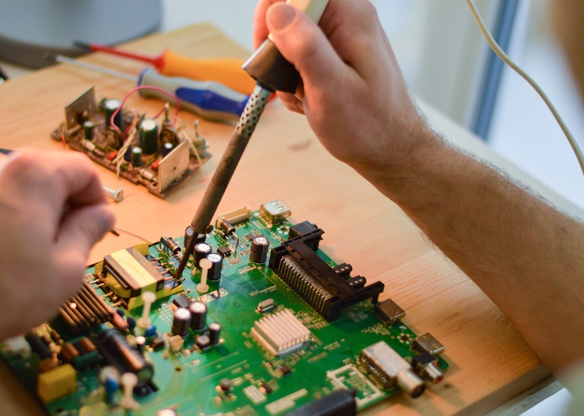 Person uses soldering iron to repair circuit board, working with small components and tools on wooden desk.