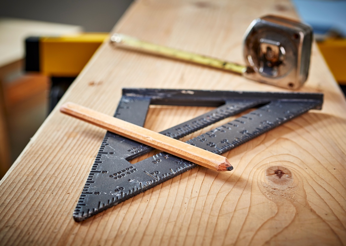 A pencil, carpenter's square, and a roll of measuring tape all sitting on top of a long piece of wood.