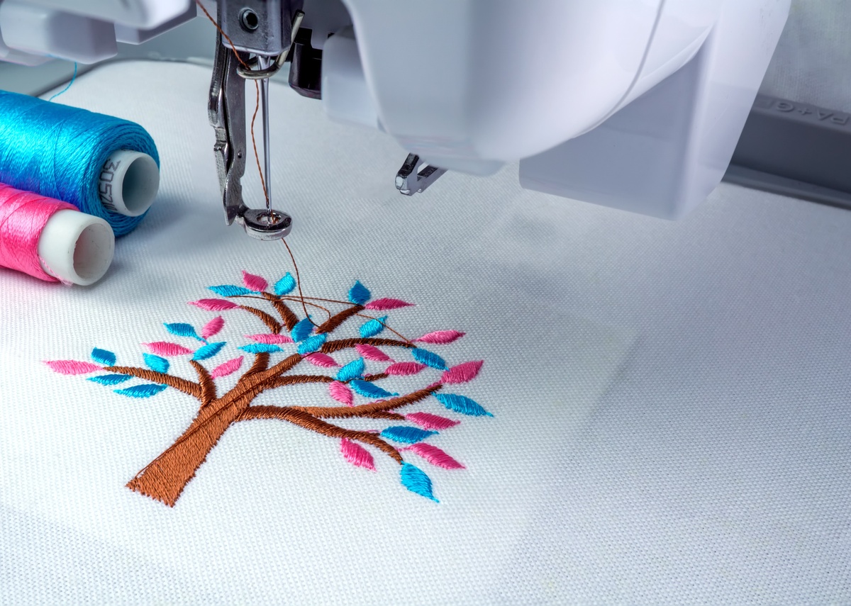 A close-up of an embroidery machine stitching a colorful tree design on white fabric with blue and pink thread spools nearby.