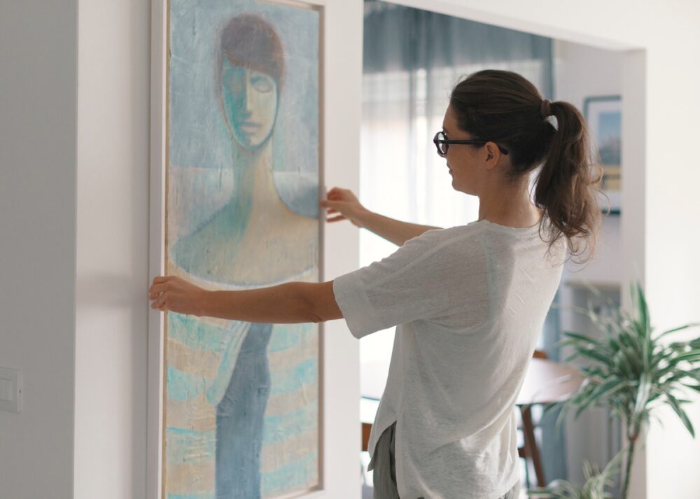 A person adjusts a large, framed painting on a wall. The artwork depicts an abstract figure in blue, beige, and green.