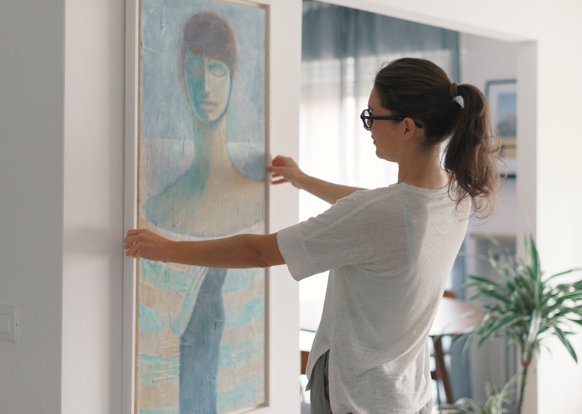 A person adjusts a large, framed painting on a wall. The artwork depicts an abstract figure in blue, beige, and green.