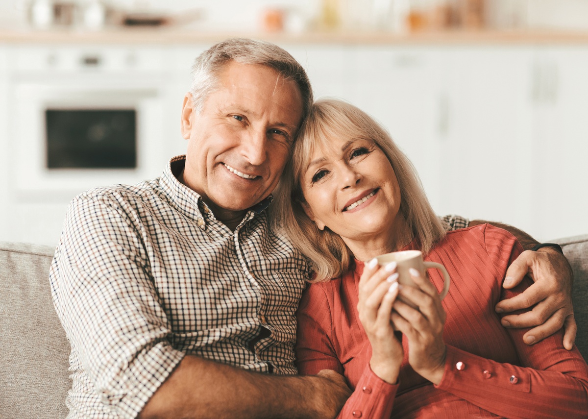 A happy boomer couple sits together on the couch cuddled into each other and smiling. The woman holds a mug.