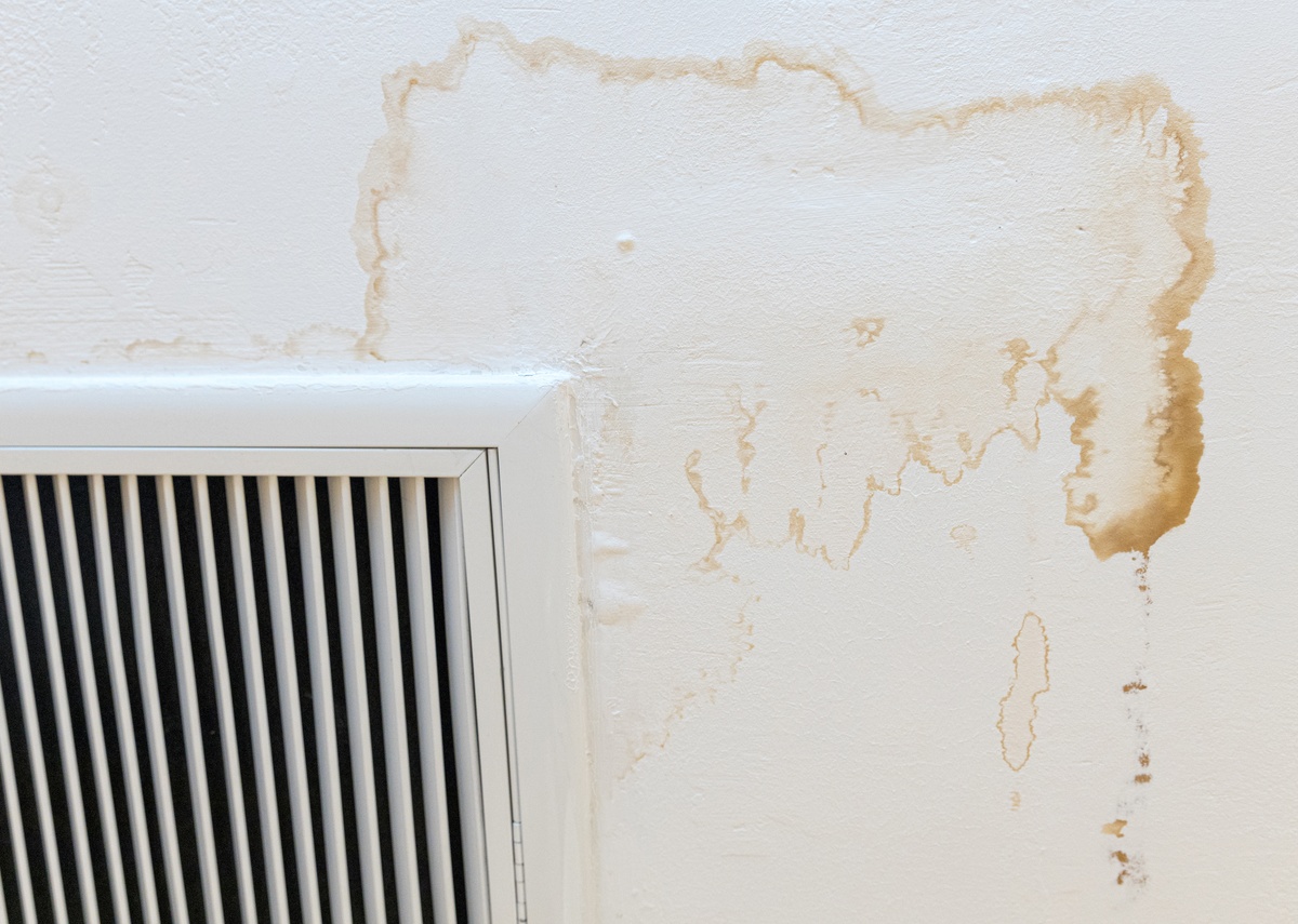 Brown water stains spread across a white wall above a vent, with drip marks and peeling texture near the corner.