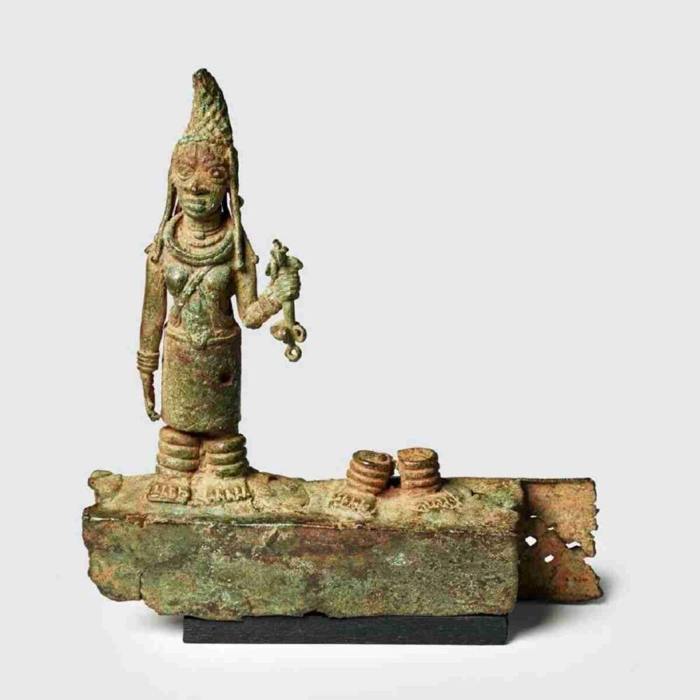 Switzerland Returns Benin Bronzes to Nigeria: An altar fragment depicting a Benin woman dressed in traditional regalia, wearing an elaborately decorated hat, holding a ceremonial staff, adorned with coral beads around her neck and heavy ivory anklets.