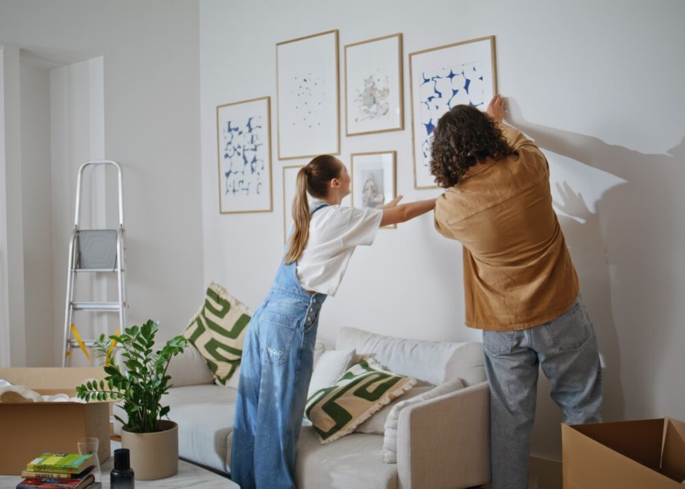 A man and woman standing by a white wall while hanging framed pictures above a sofa in a bright living room. They are using tips for decorating your home with art and collectibles