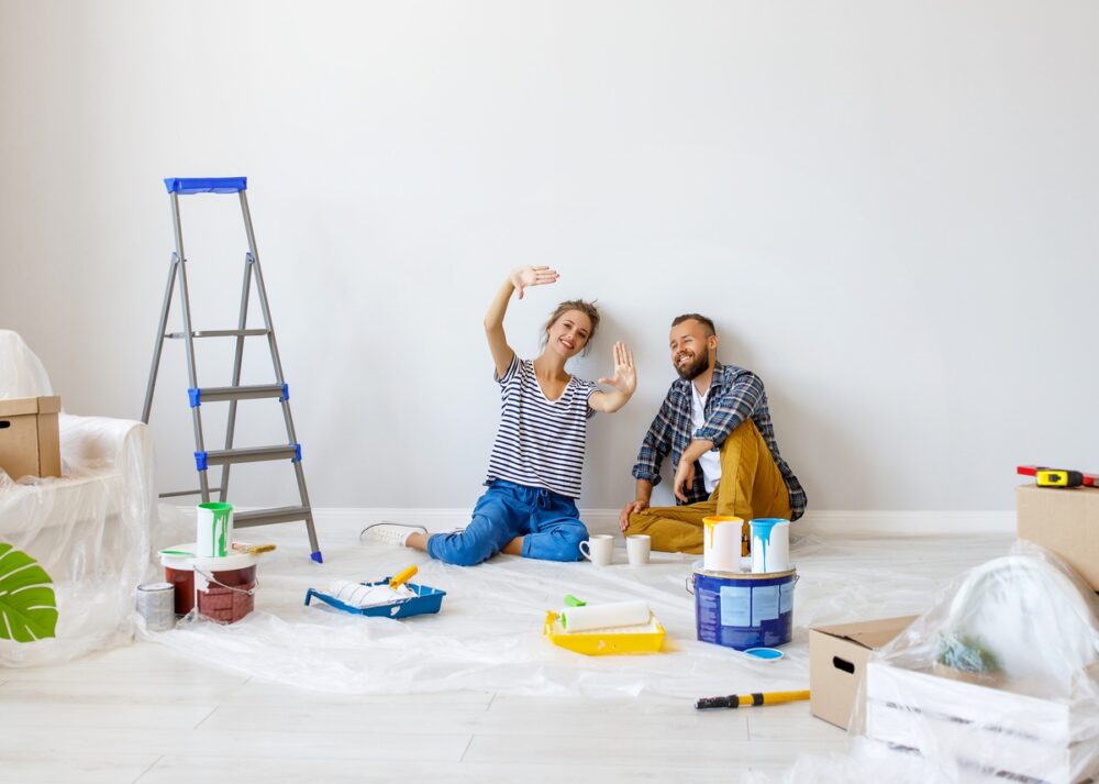 A couple sits on the floor in their renovated living room. One person holds their hands to envision a window.